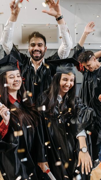 Graduates in black robes celebrate by tossing confetti in the air, smiling, and holding diplomas.