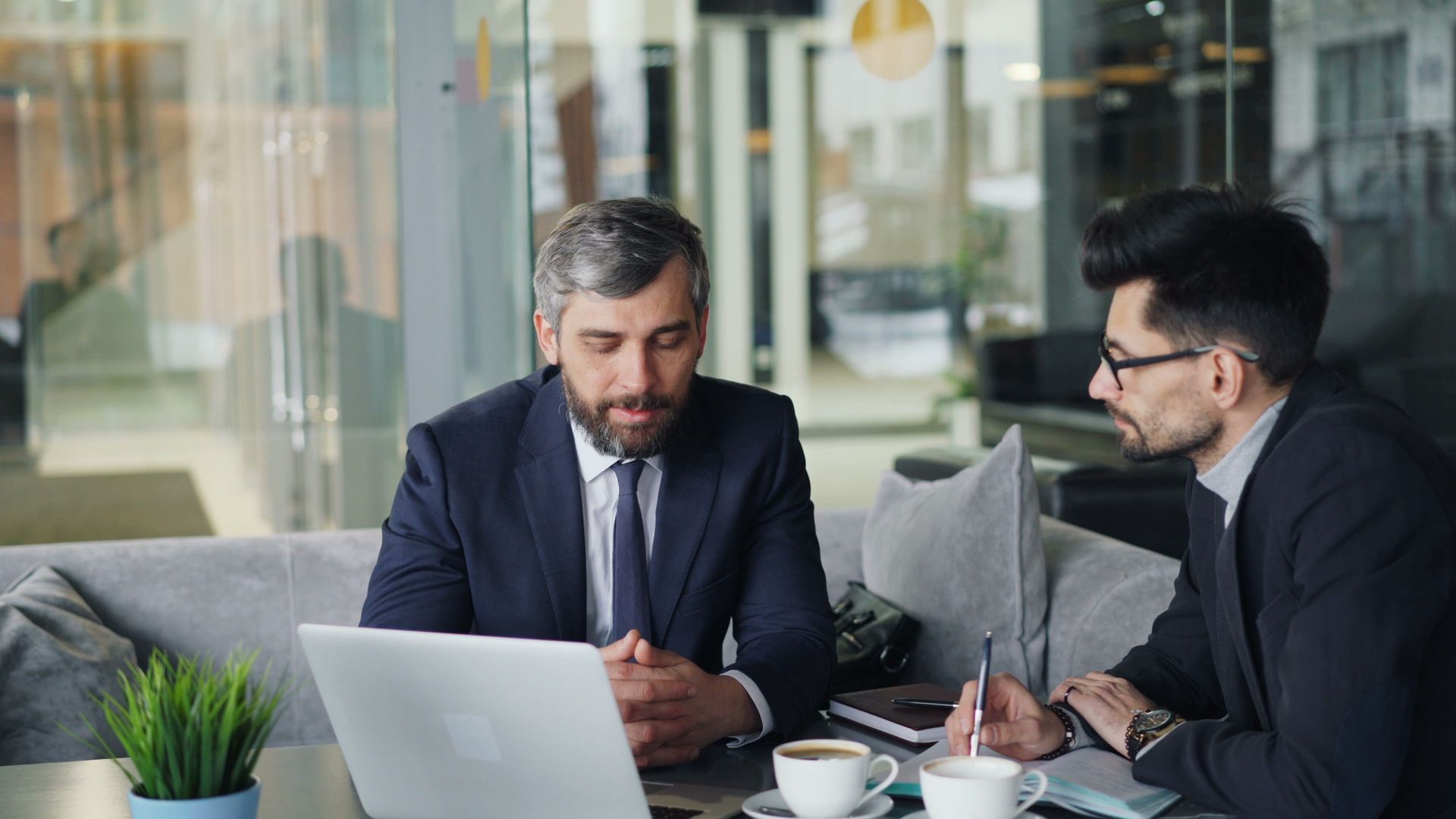 Two men in suits discussing work at a table with a laptop in a modern office lounge.