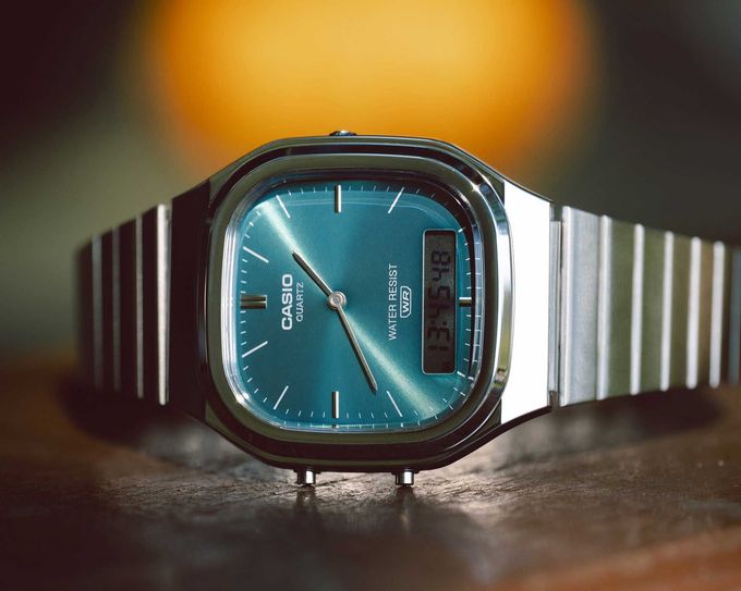 Un orologio da polso Casio ana-digi color argento con quadrante a raggiera color verde acqua, appoggiato su una superficie scura contro uno sfondo caldo e sfocato.