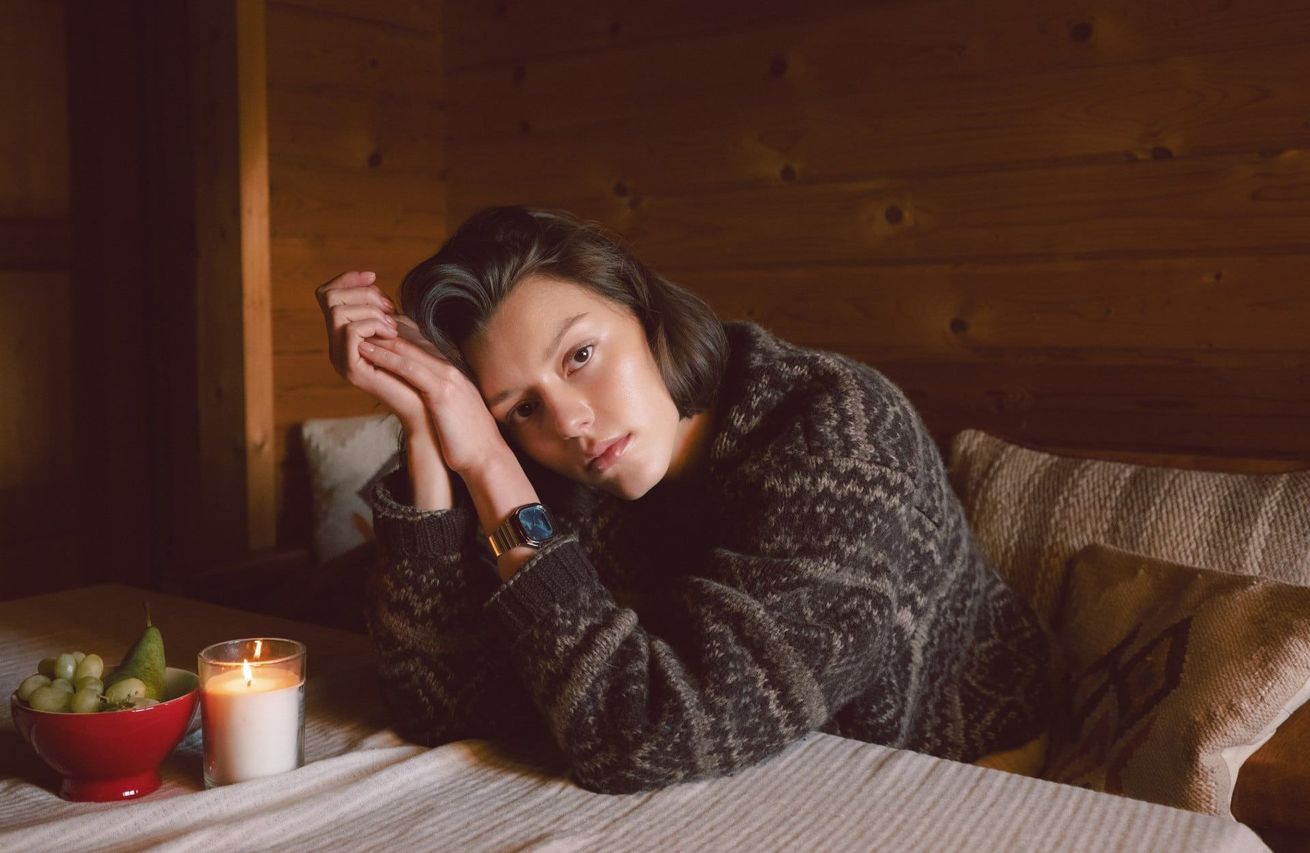 In una stanza, una persona dai capelli scuri indossa un maglione a fantasia ed è appoggiata a un tavolo accanto a una candela e a una ciotola d'uva.