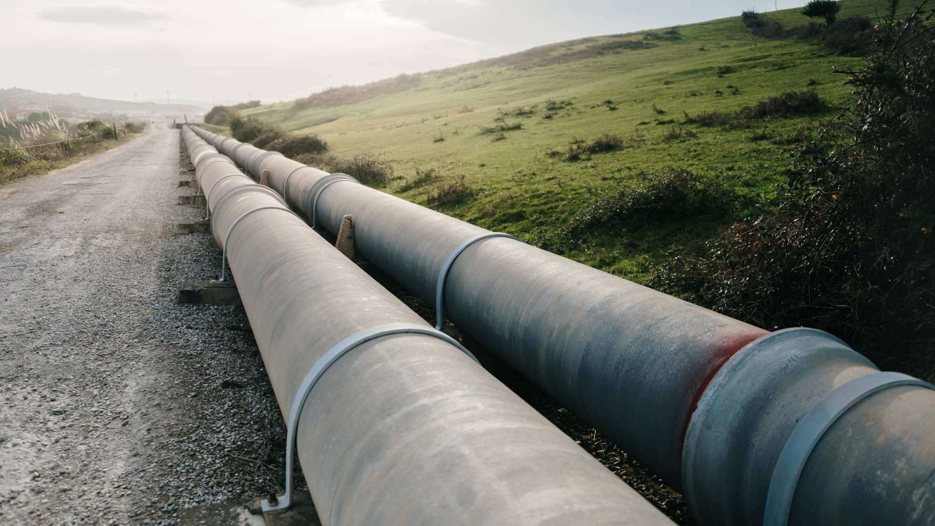 Large pipeline runs along a gravel road through a grassy hillside landscape.