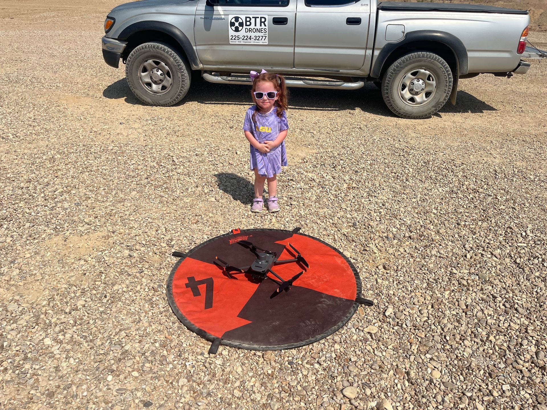 Child standing on gravel near a red-and-black circular object, with a silver pickup truck behind.