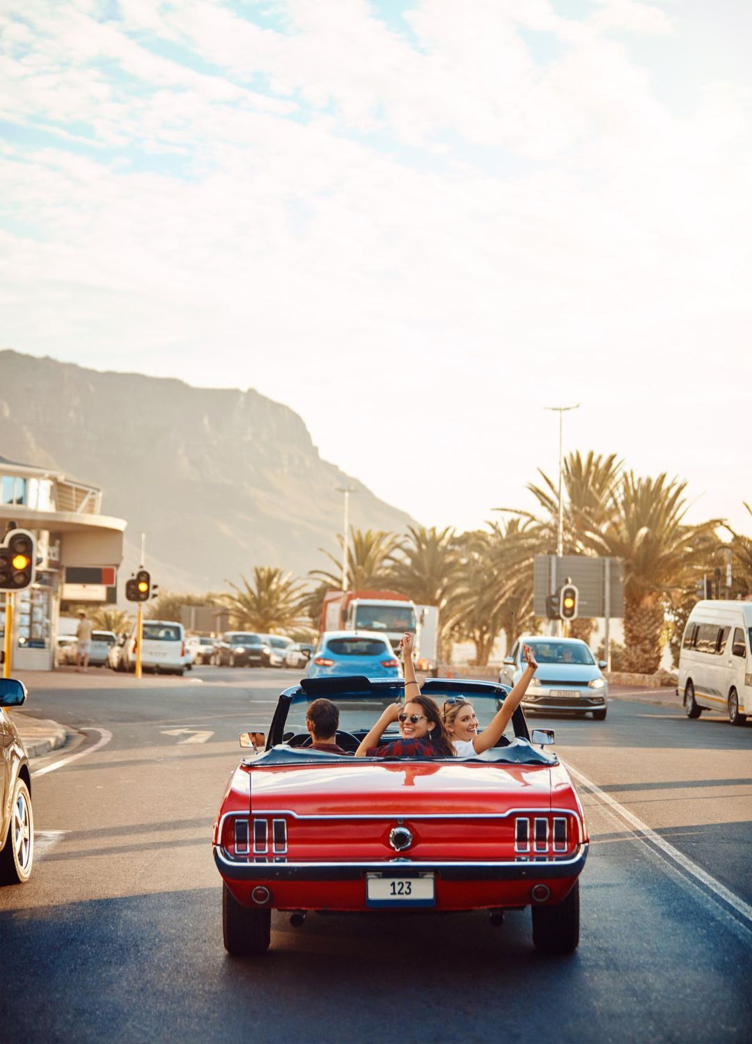 Three people in a bright red convertible cruising on a sunlit coastal road with palm trees and a mountain in the background.