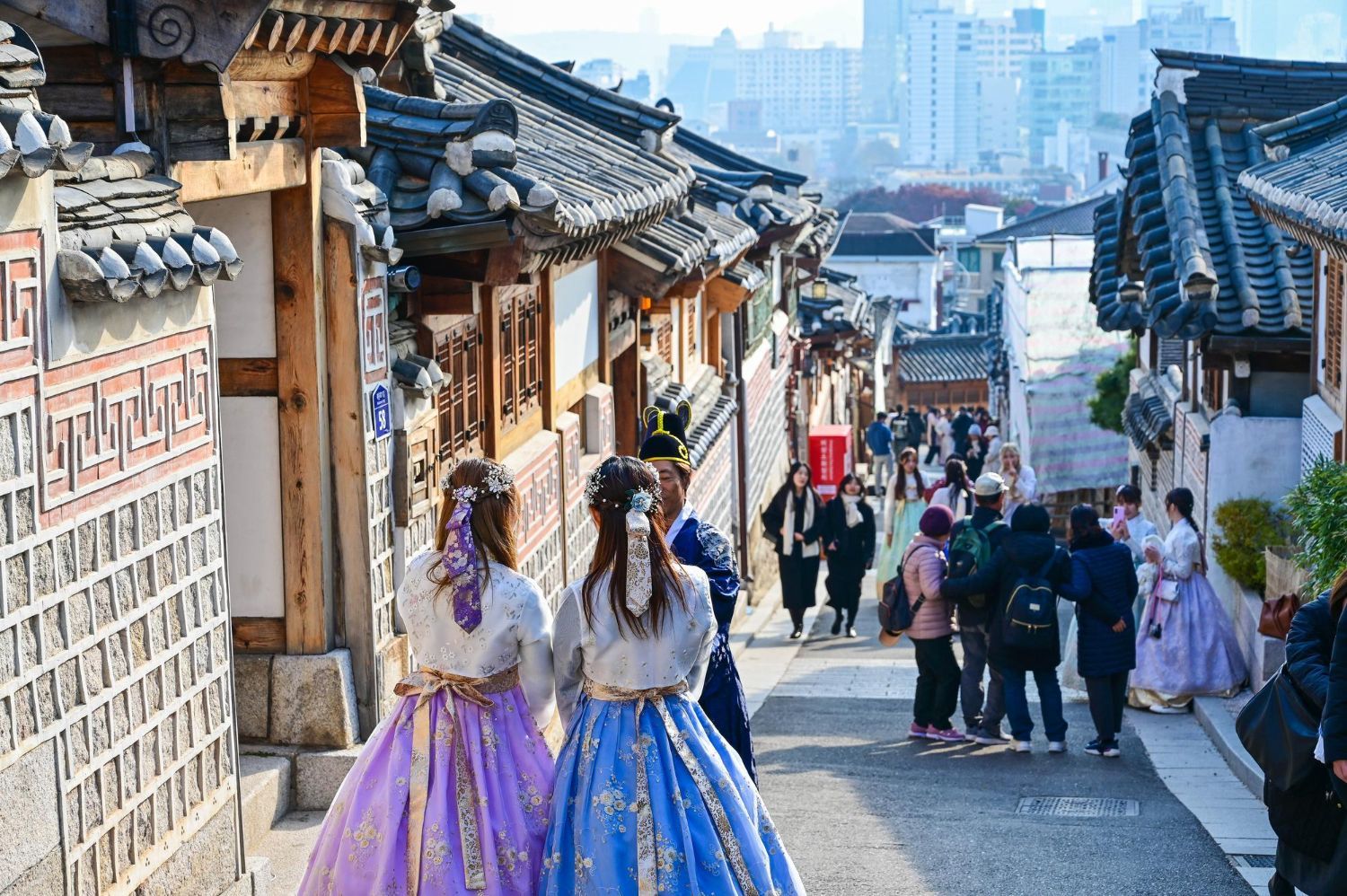 People in traditional Korean hanbok stroll down a narrow alleyway lined with historic tiled-roof houses in Seoul.