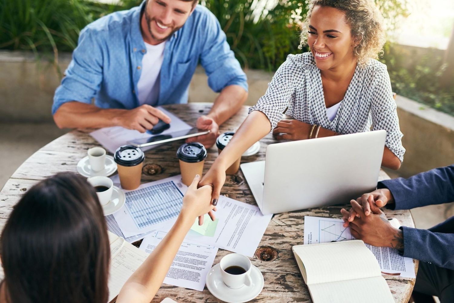 Four people sit around a wooden table with laptops and documents as two reach across to shake hands during a meeting.