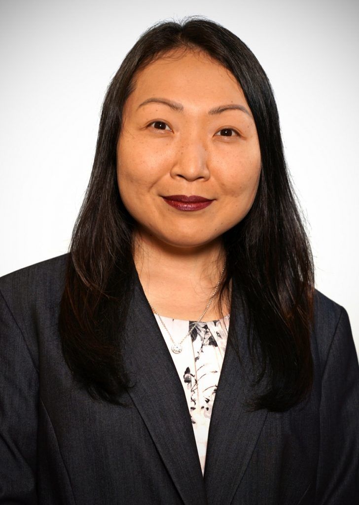 A professional headshot of a person with long dark hair, wearing a dark blazer over a patterned shirt against a light wall.