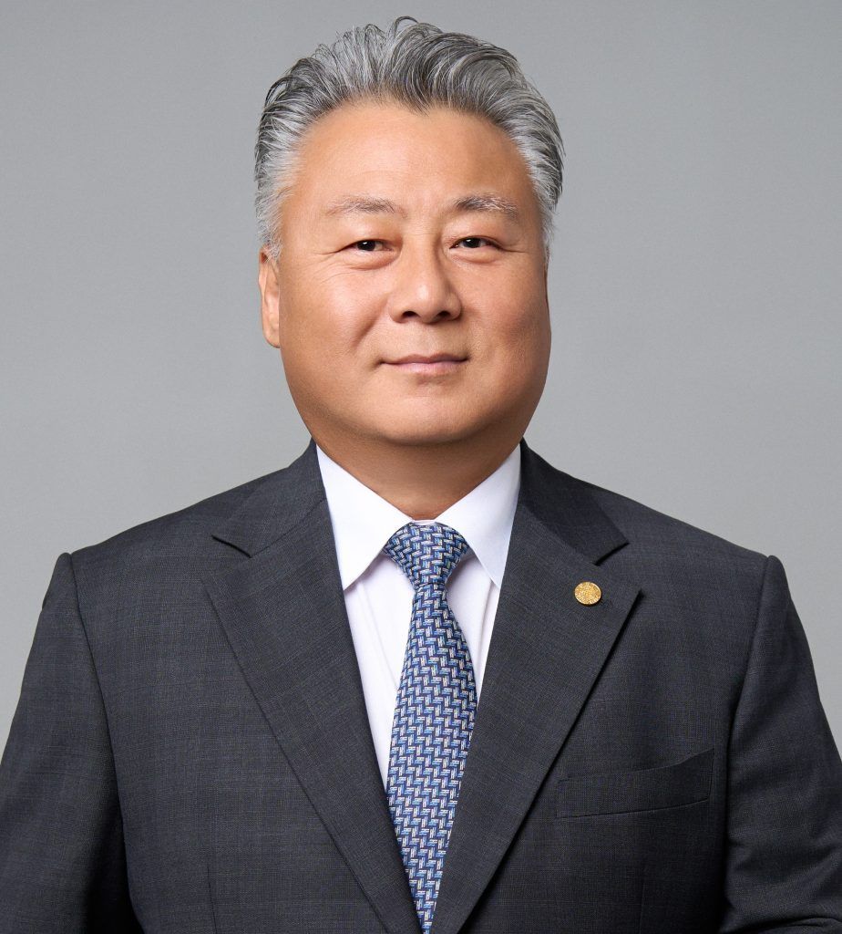 A professional headshot of a person in a grey suit, white shirt, and patterned blue tie, posed against a grey background.