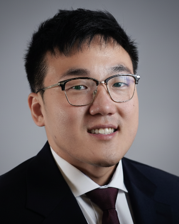 A headshot of a person wearing glasses, a dark suit, and a maroon tie against a plain gray background.