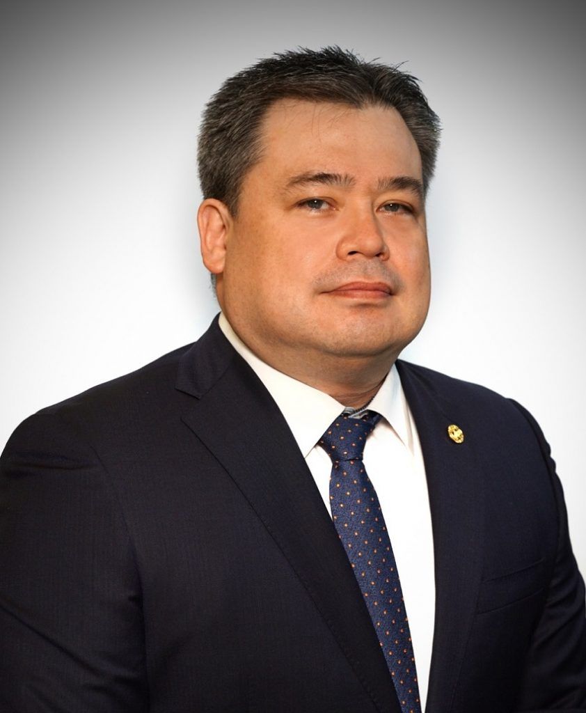 Headshot of a person in a navy blue suit, white collared shirt, and dark blue patterned tie against a light background.