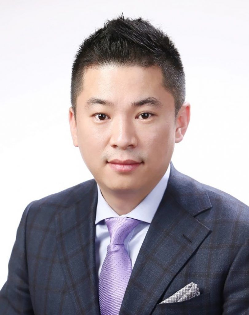 A professional headshot of a person in a dark plaid suit, light blue shirt, and purple tie against a white background.