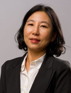 A professional headshot of a person with shoulder-length dark hair, wearing a white collared shirt and black blazer.