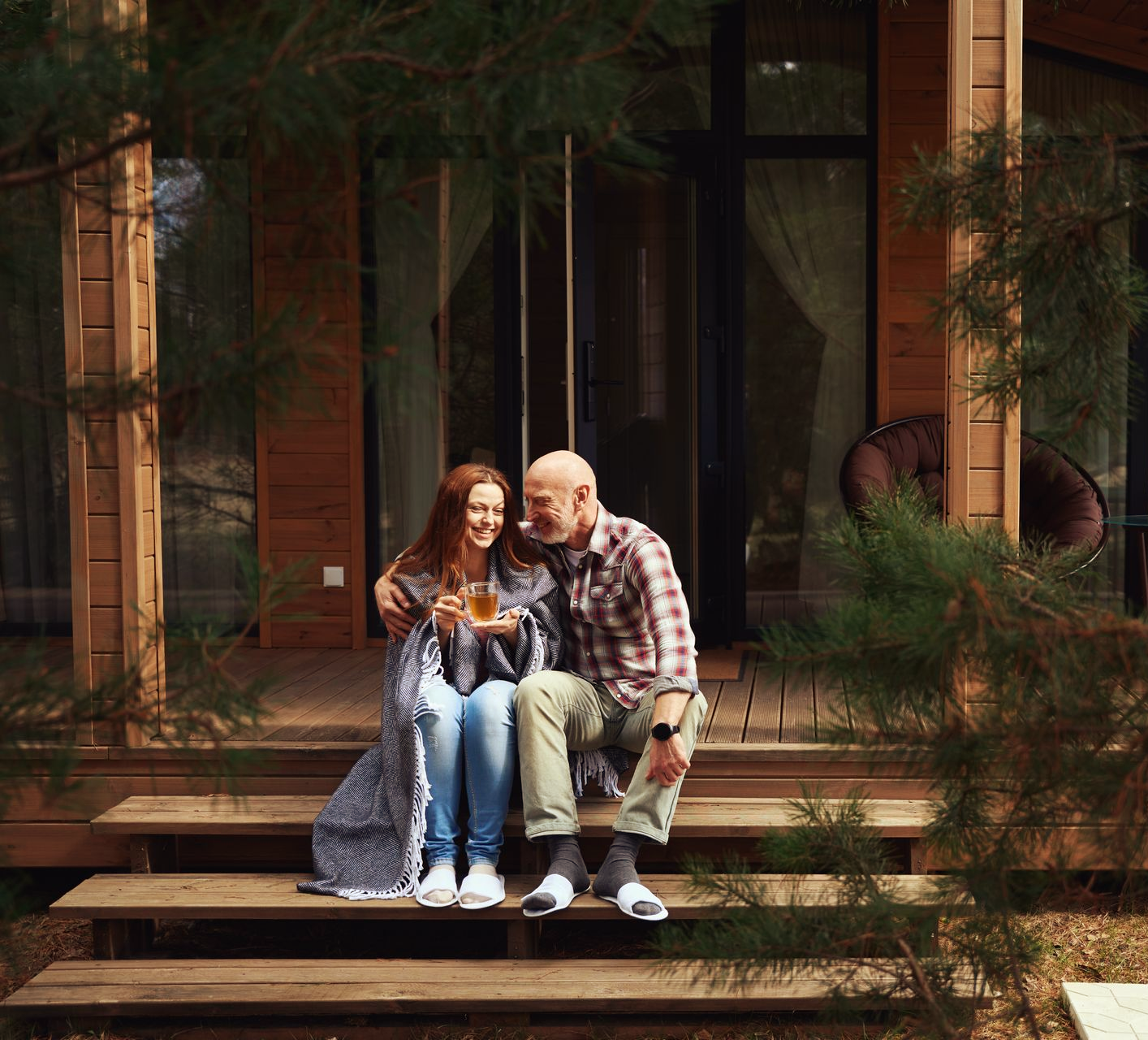 A person wrapped in a blanket and an individual in a plaid shirt sit on wooden porch steps, holding cups and smiling.