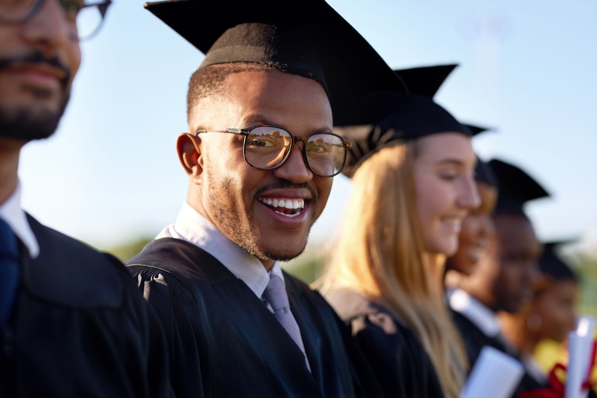 A group of graduates in caps and gowns stand in a line, with one person in the foreground smiling.