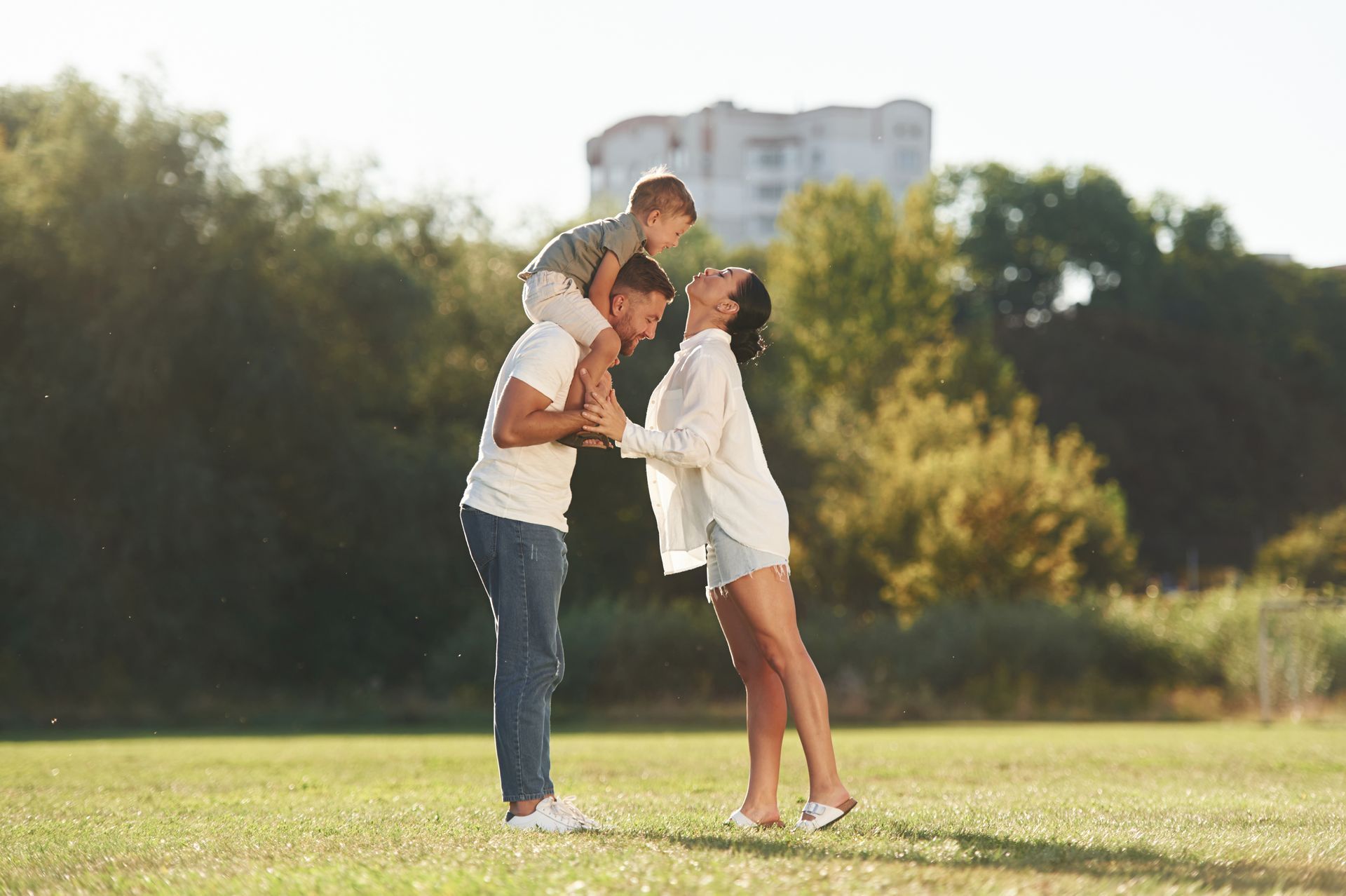 A family of three standing in a sunny park, with a child sitting on a parent's shoulders as they face each other.