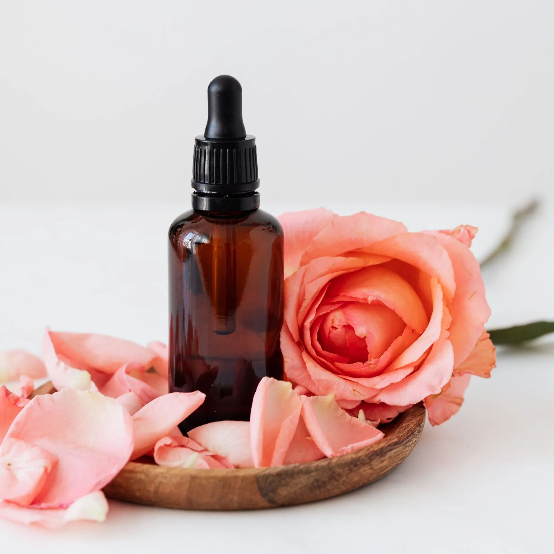 A bottle of essential oil next to a pink rose on a wooden plate.