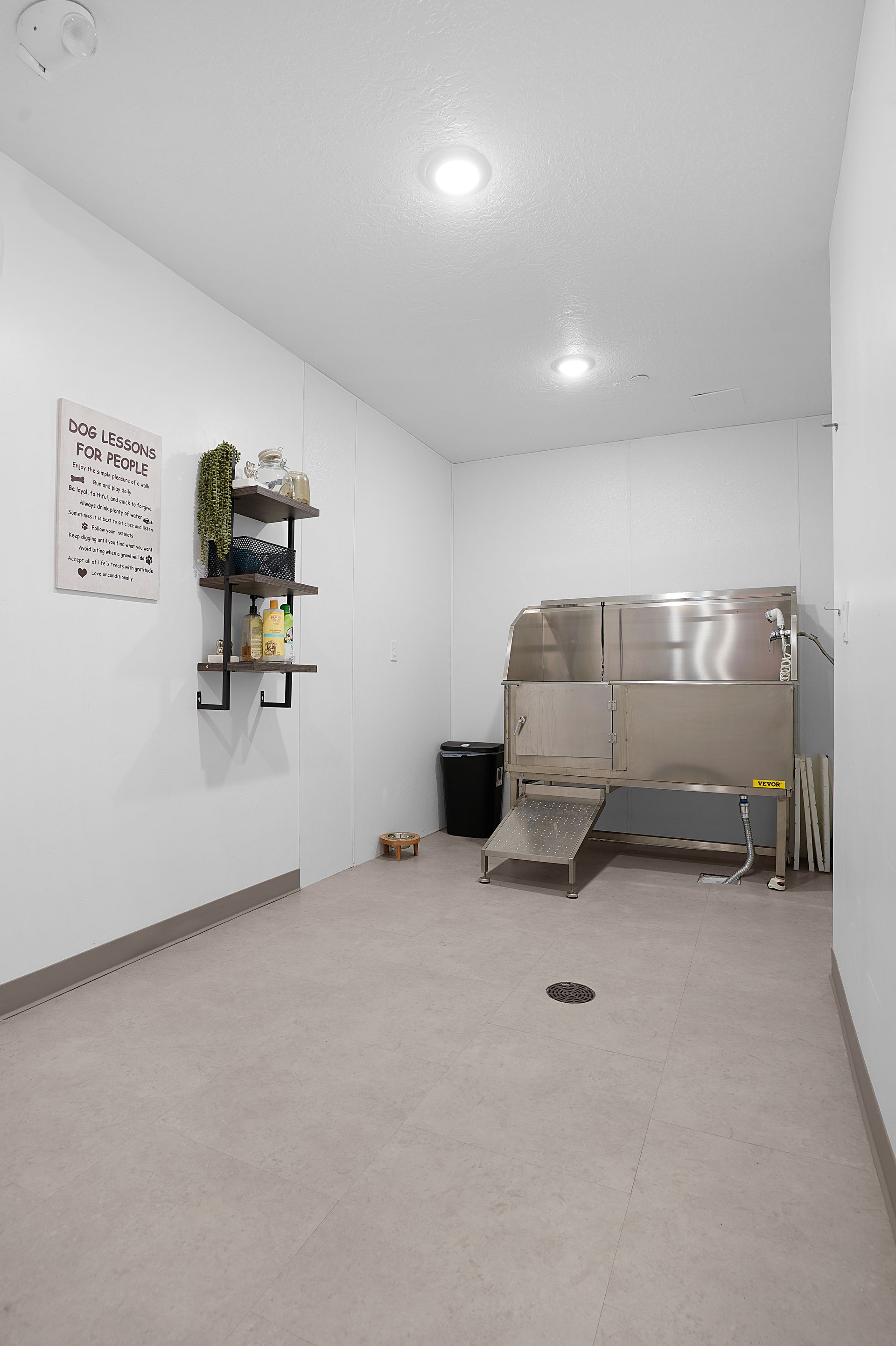 A dog grooming room with stainless steel equipment, shelves with products, and a trash can at Sorella in Herriman, UT.