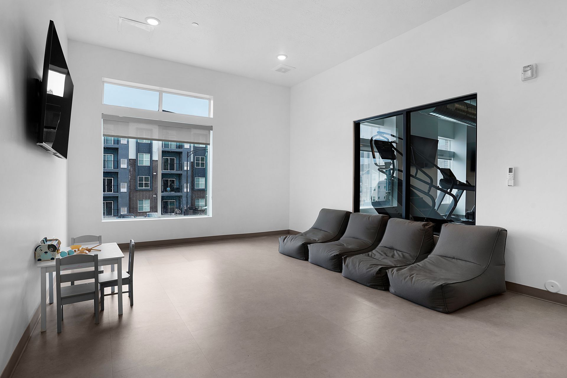 Modern room with beanbag chairs, a small table, a window, and a television. A gym can be seen through a glass door at Sorella in Herriman, UT.