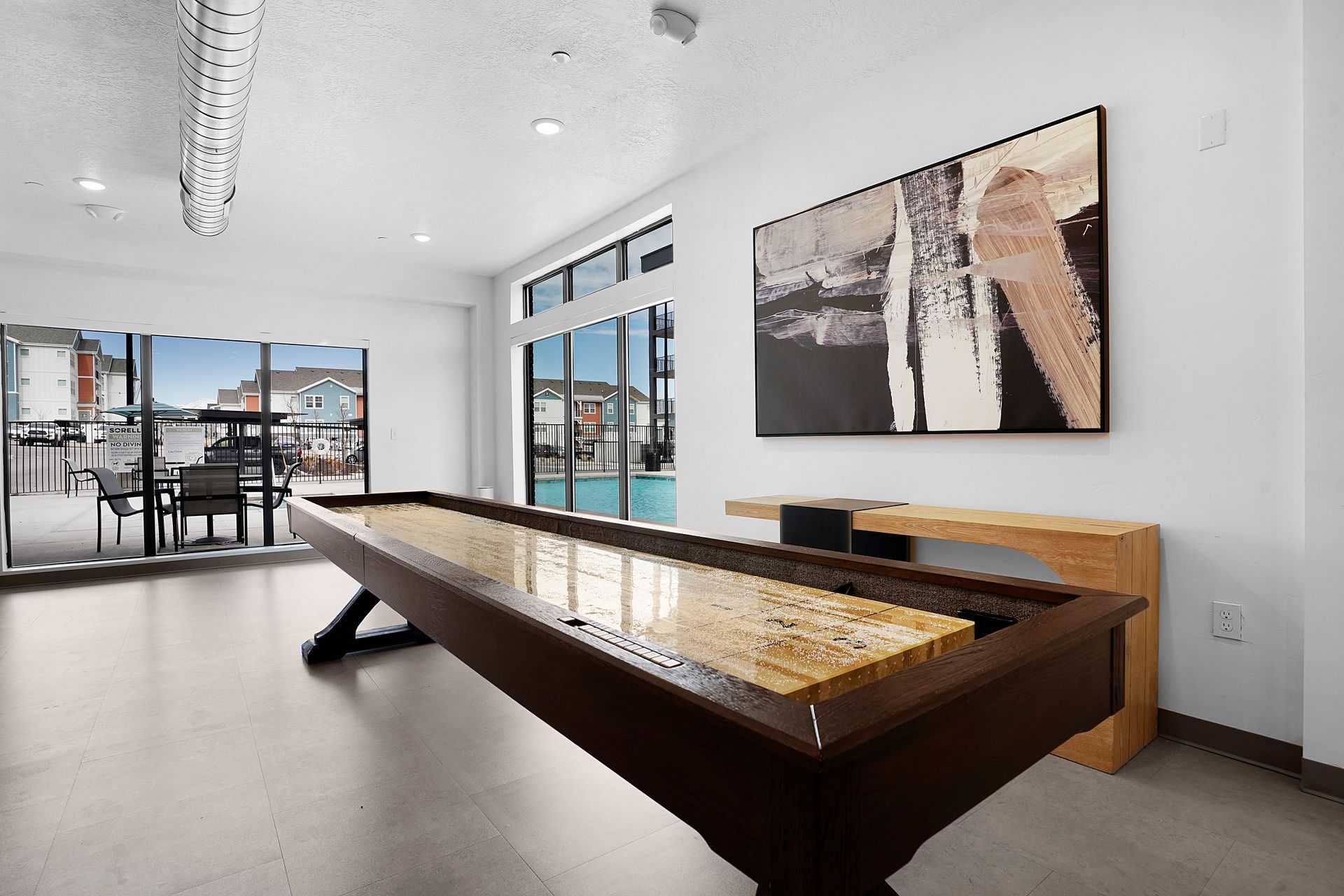 A shuffleboard table in a room with large windows overlooking an outdoor area with tables and a pool at Sorella in Herriman, UT.