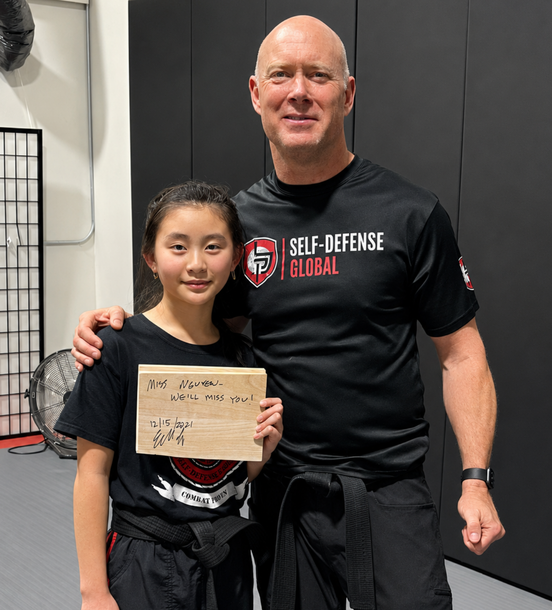 Two people in a gym hold a certificate, both in black Self-Defense Global shirts.