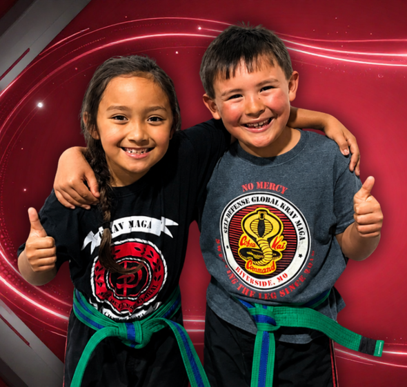 Two smiling children in martial arts uniforms with green belts, standing arm-in-arm against a red swirled backdrop