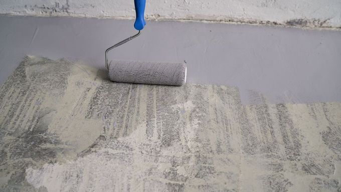 Paint roller applying white paint to a gray wall/floor surface.