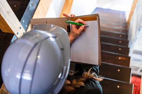 Worker in hard hat writing on a clipboard beside an indoor stairwell