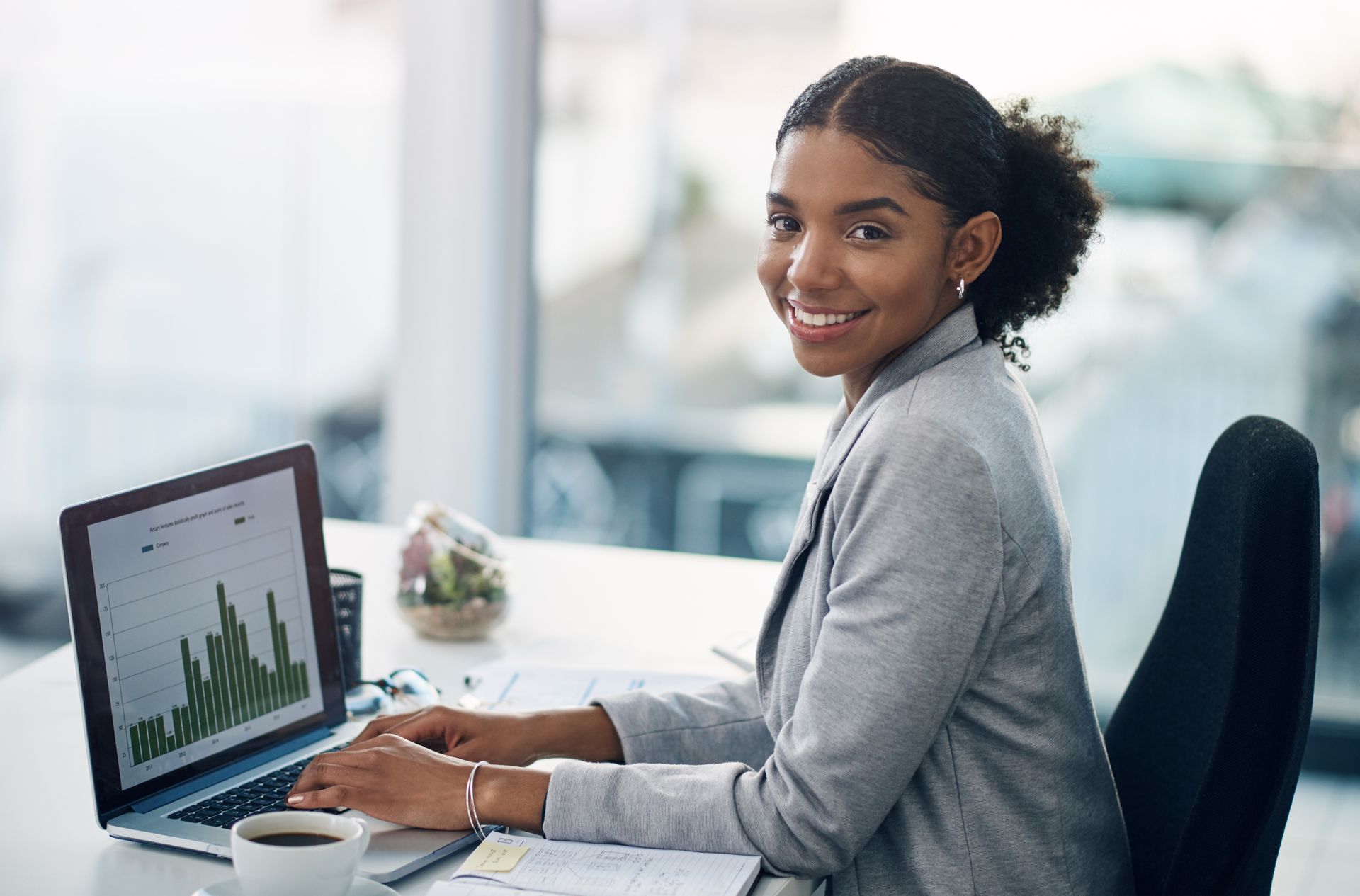 Smiling professional at desk using laptop with analytics charts in a bright office