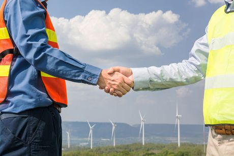 Two people in safety vests shaking hands with wind turbines in the background