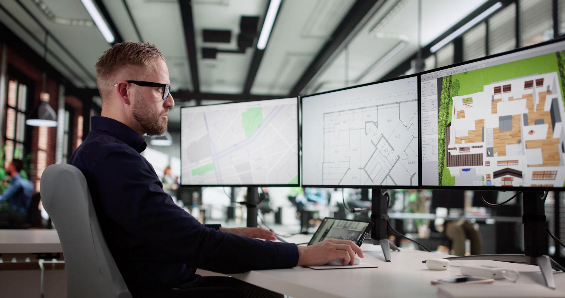 Person working at a desk with three monitors displaying maps and diagrams in a modern office