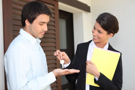 Two people at a doorway exchange keys, one holding a yellow folder and smiling