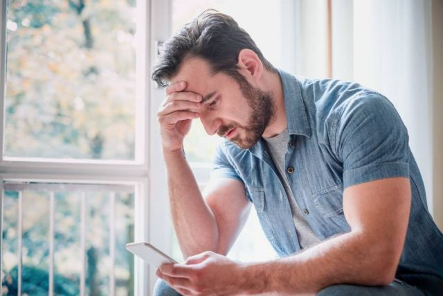 A man is sitting on a window sill looking at his cell phone.