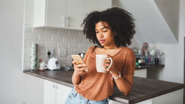 A woman is standing in a kitchen holding a cup of coffee and looking at her phone.