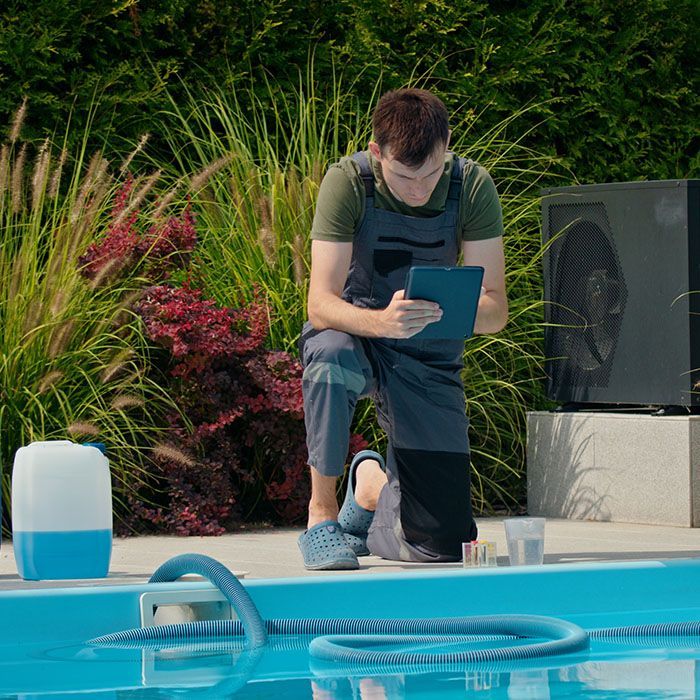 Person kneels by a pool, reading a blue clipboard beside a vacuum hose and landscaping.