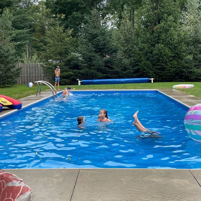 Backyard pool with children swimming and playing, surrounded by trees and pool toys.