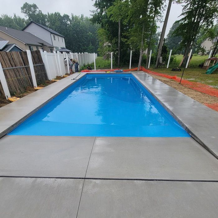 Rectangular outdoor swimming pool with bright blue water and a concrete deck, surrounded by trees and houses
