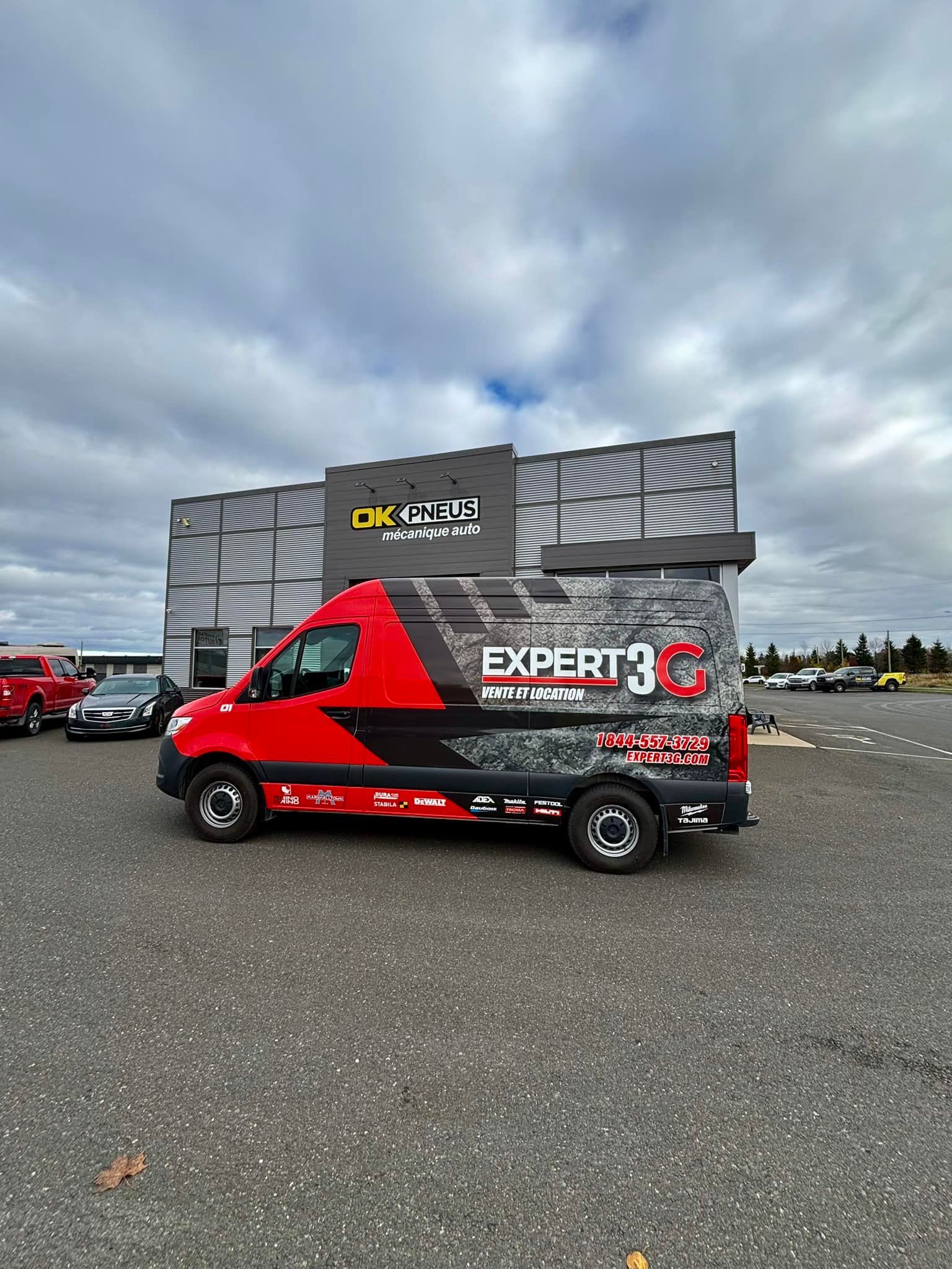 Une camionnette rouge et noire de marque Expert est garée sur du gravier devant un bâtiment sous un ciel nuageux.