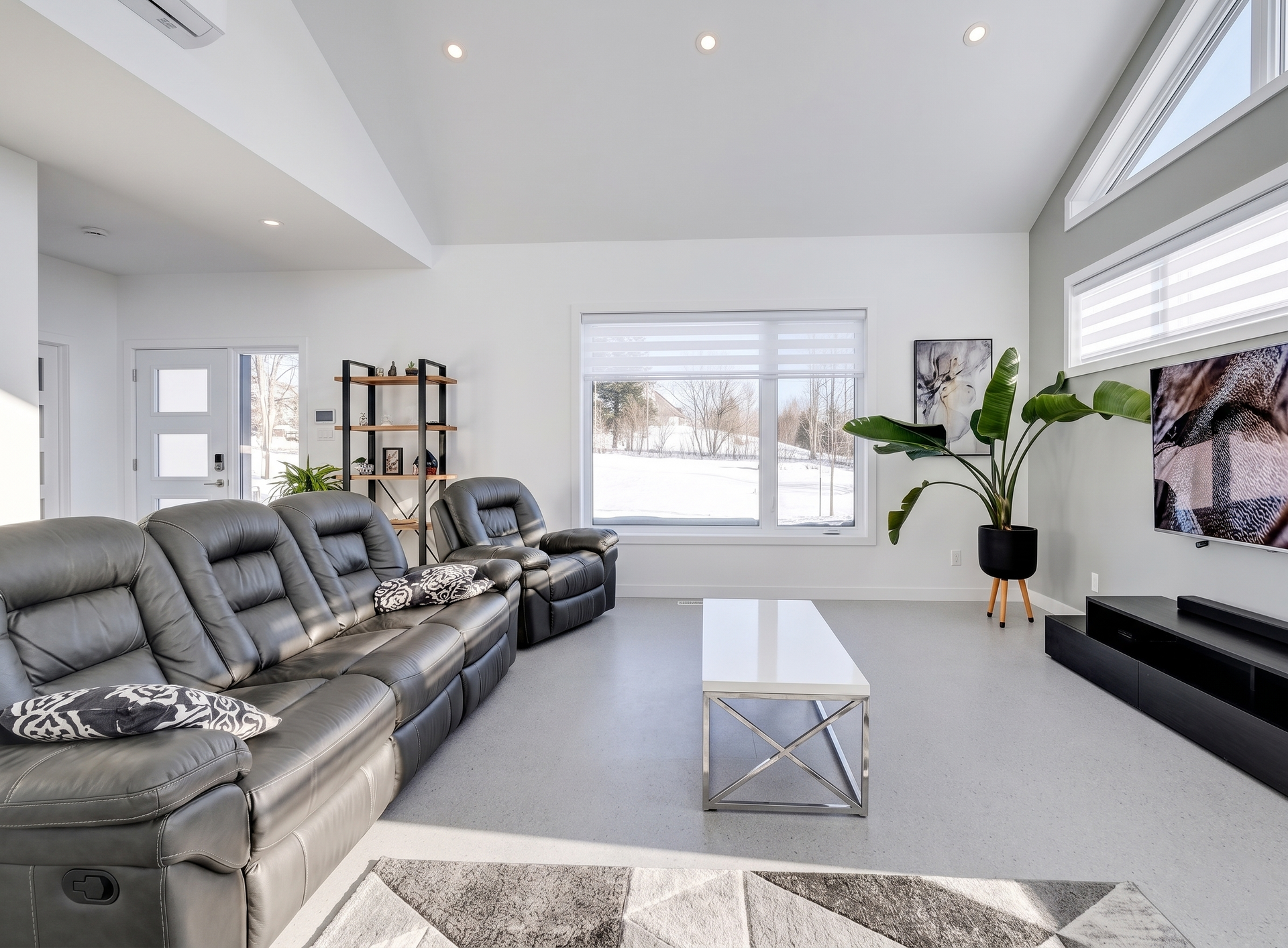 Salon moderne et lumineux avec canapé d'angle gris, table basse blanche et grandes fenêtres donnant sur l'extérieur