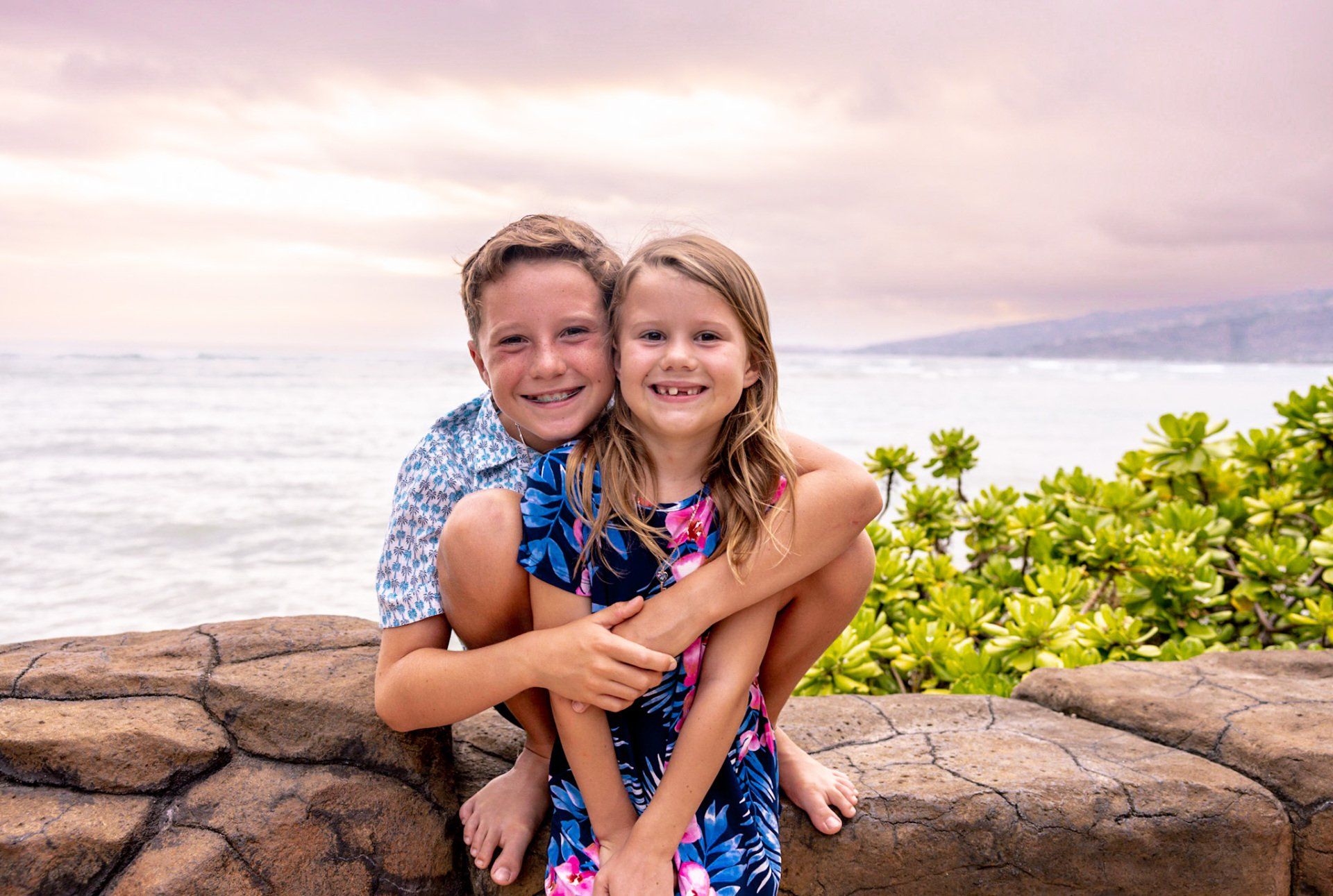 A boy and a girl are sitting on a rock wall near the ocean.