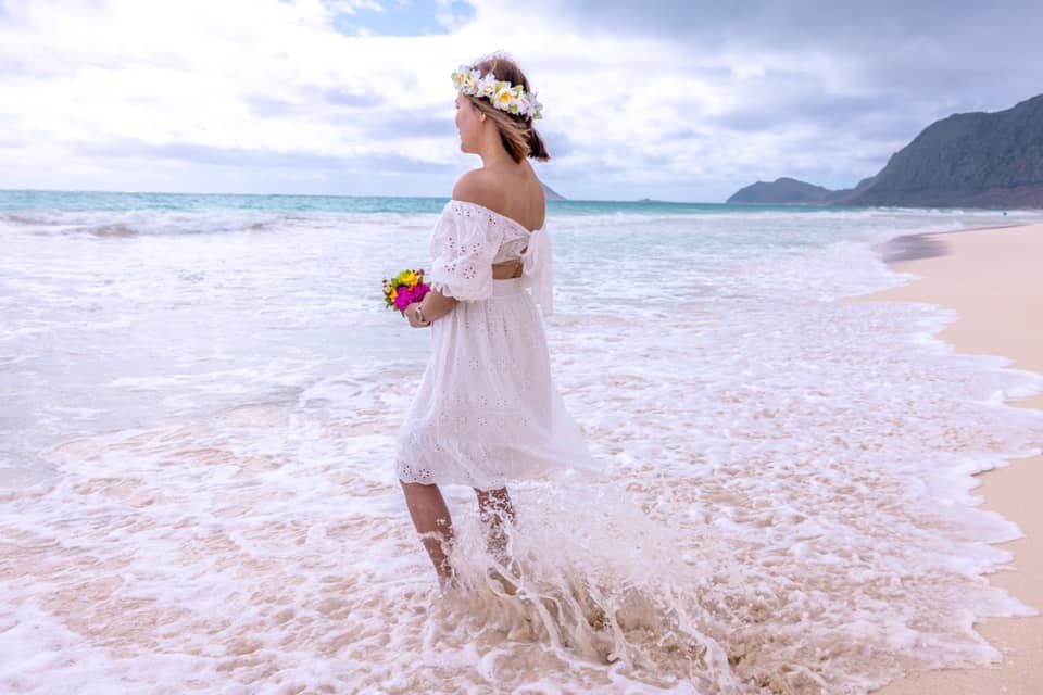 A woman in a white dress is standing on a beach holding a bouquet of flowers.