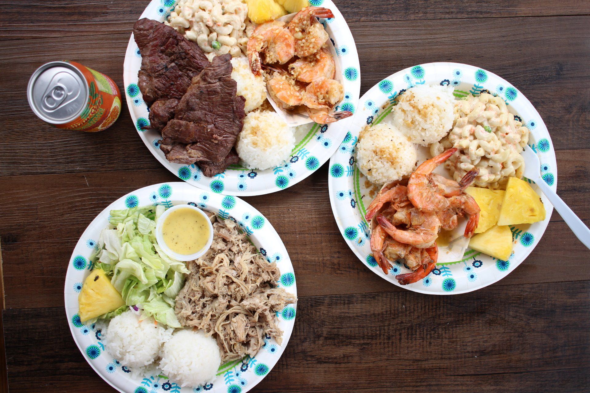 Three plates of food and a can of soda on a wooden table.