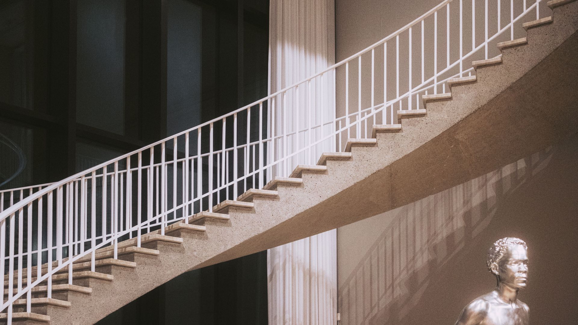 Diagonal white staircase with railing against a shadowed building; small statue at lower right.