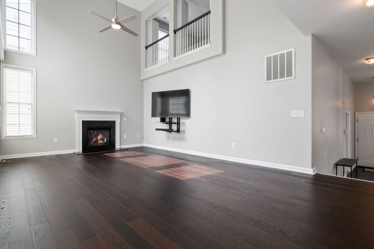 Empty modern living room with dark wood floors, gray walls, fireplace, wall-mounted TV, and stair railing above