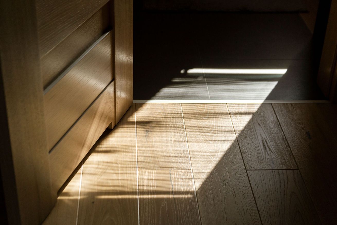 Sunlight streaming through blinds onto a wooden floor beside a cabinet in a dim room