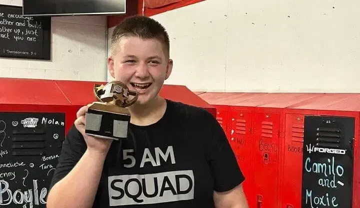A young man in a 5 am squad shirt is holding a trophy.