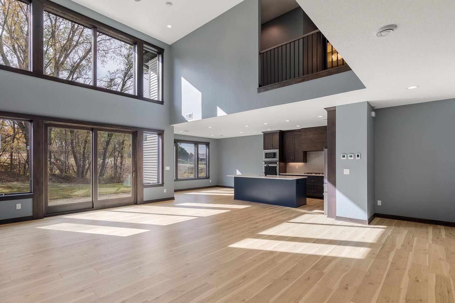 Spacious modern home interior with large windows, light wood floors, and blue-gray walls; kitchen in the back.