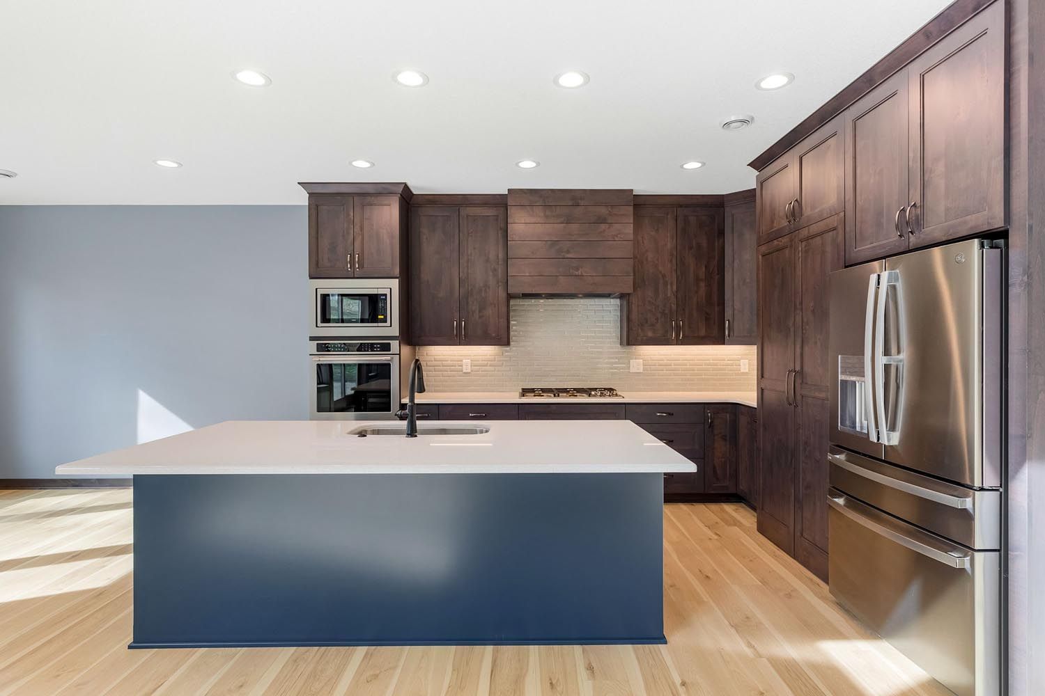 Modern kitchen with dark wood cabinets, blue island, stainless steel fridge, and light wood floors.