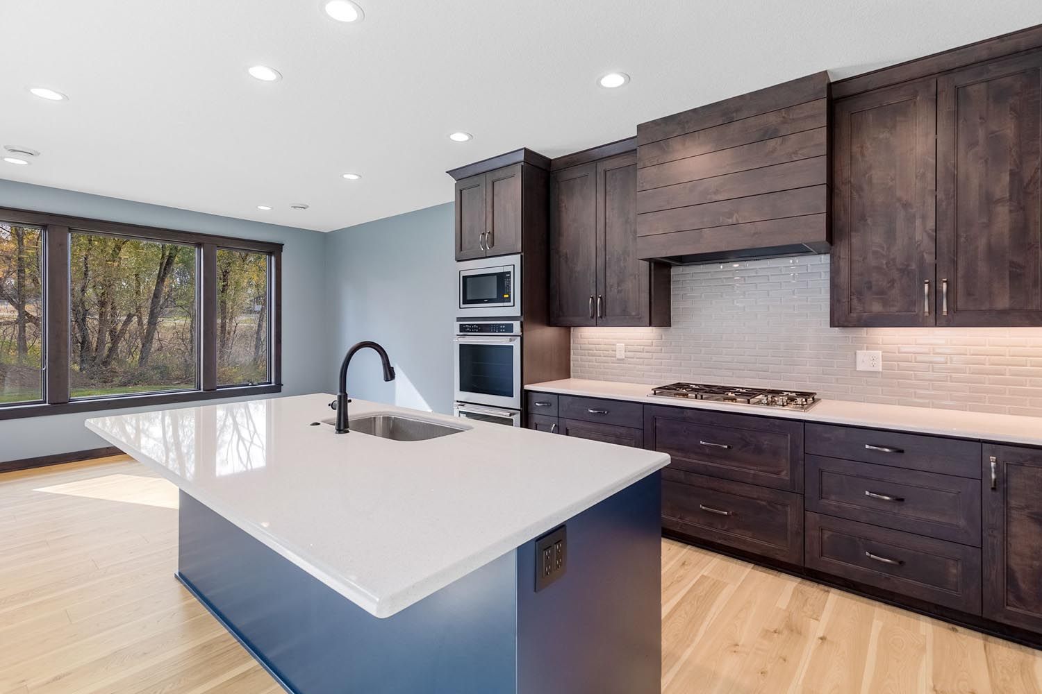 Modern kitchen with blue island, dark wood cabinets, white countertops, and large window overlooking trees.