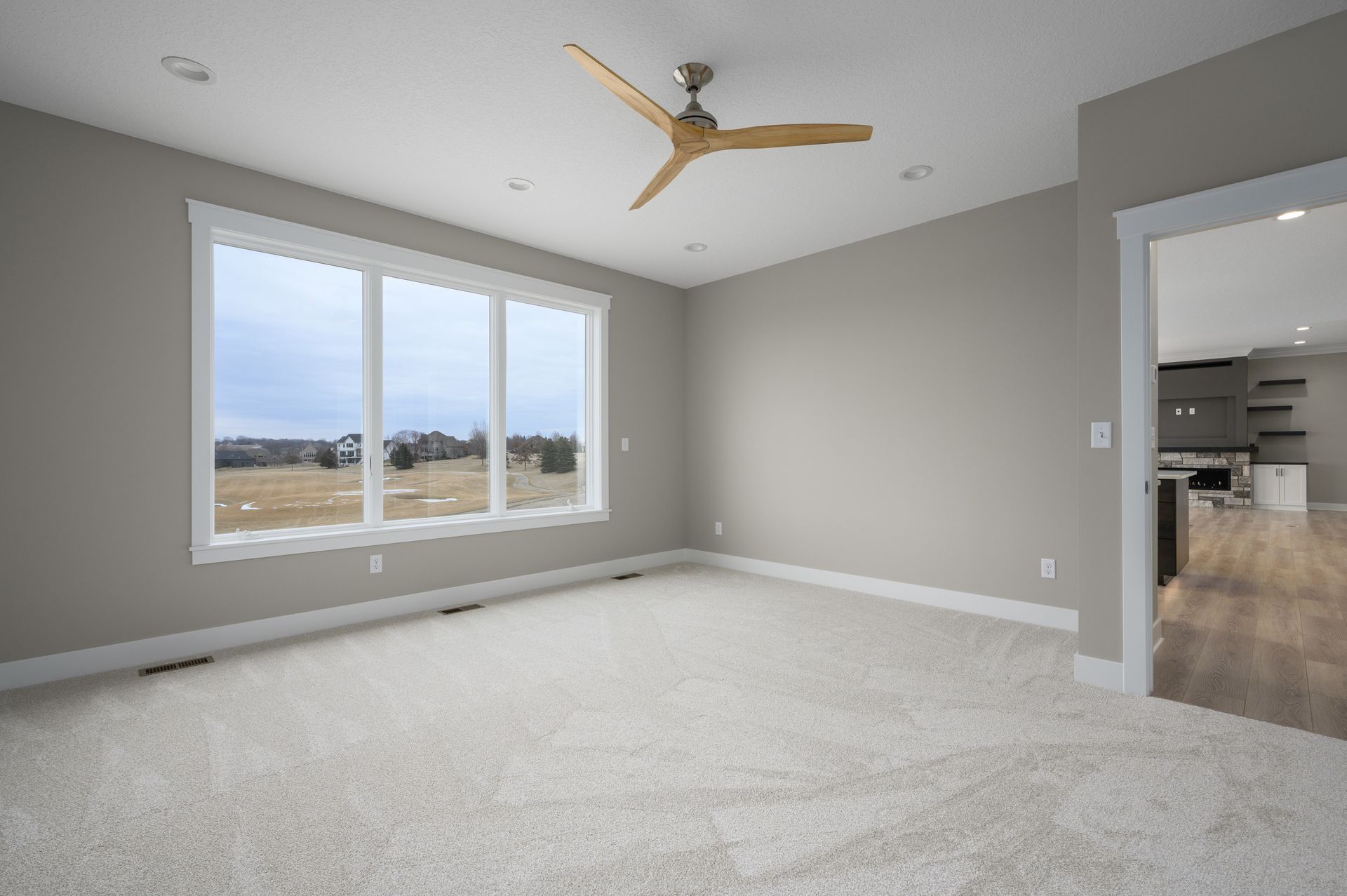 Empty bedroom with large window, light carpet, gray walls, and ceiling fan.