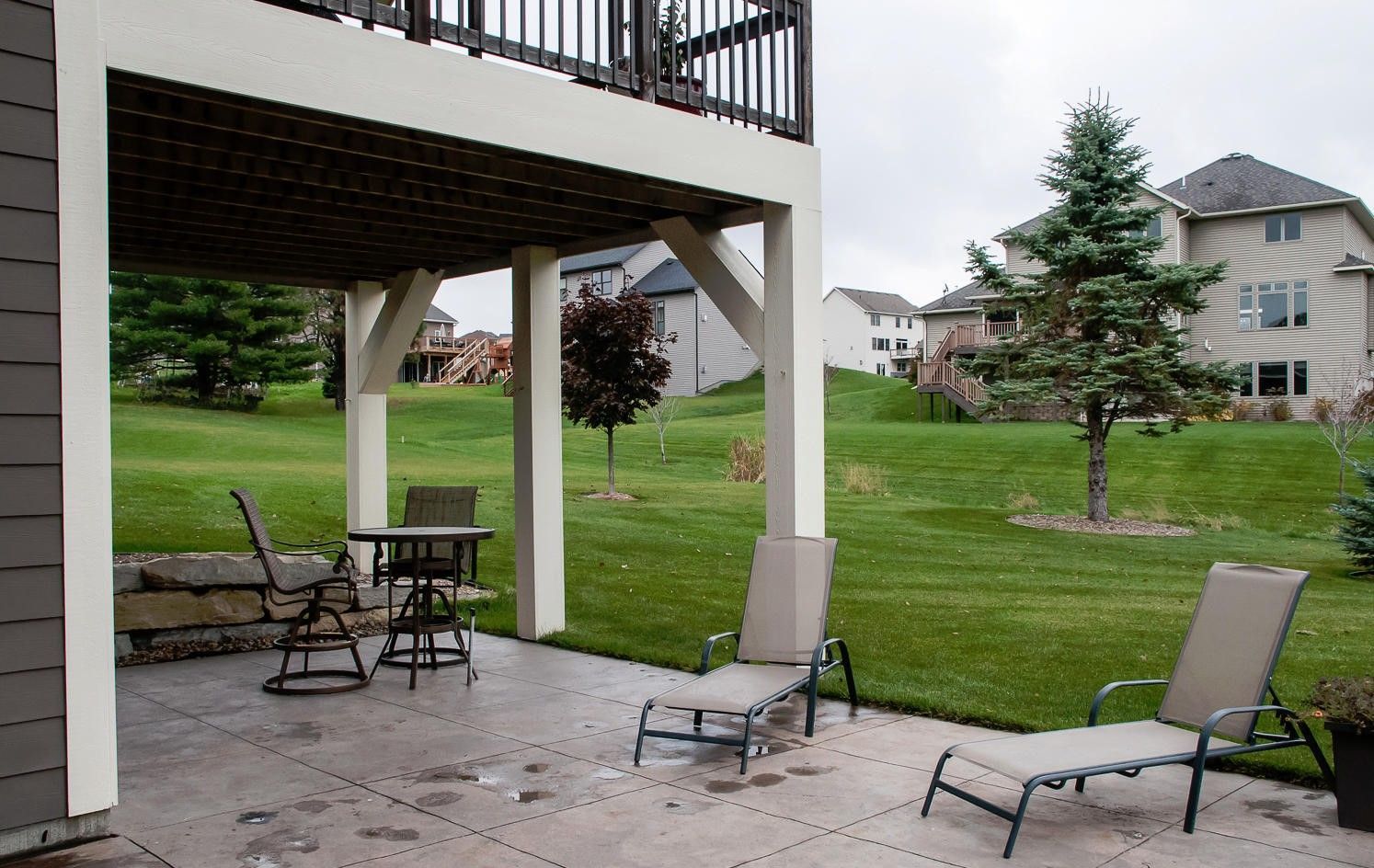 Patio under a deck with lawn chairs and a table on a concrete surface, overlooking a grassy yard with houses.
