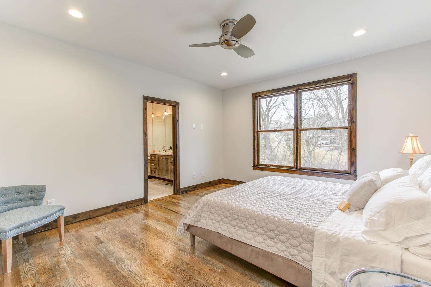 Bedroom with wooden floors and trim, a bed, a window, a chair, and a ceiling fan.